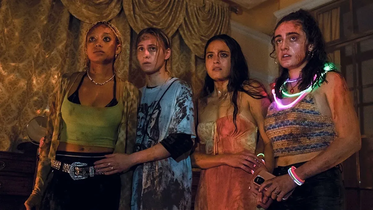 Four women stand in an expensive looking room. They are covered in rain, mud, and blood, with shocked or frightened looks on their faces.