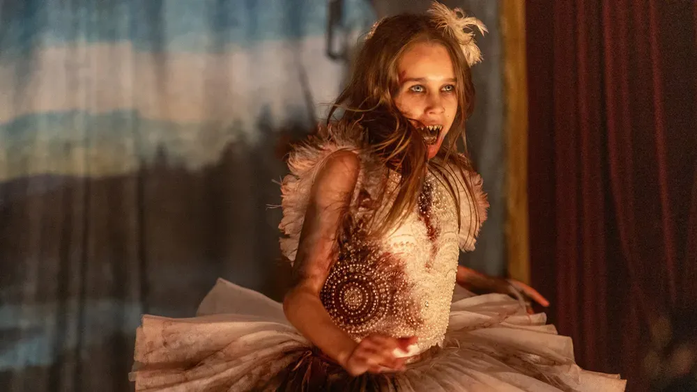 A young girl, around 12 years old, in a fancy ballerina outfit splattered with blood. She is in mid spin, revealing the her very sharp teethe as she's about to snarl.