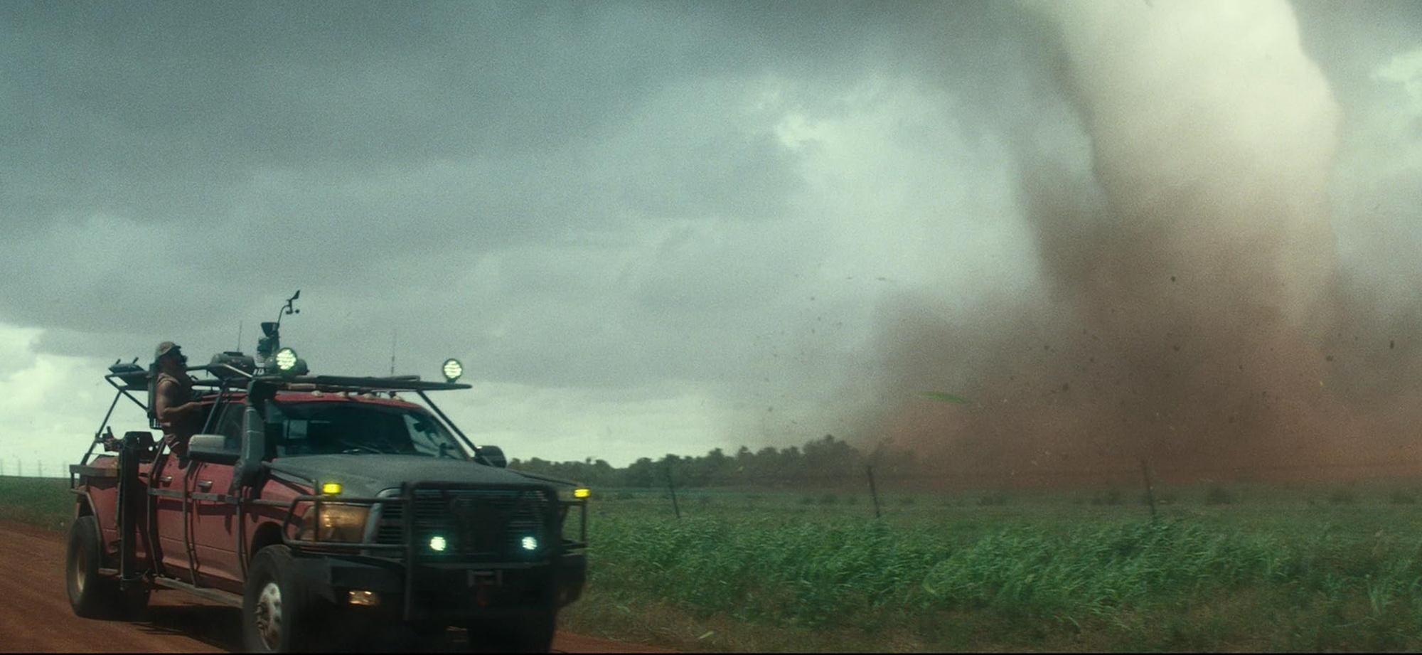 A rugged red truck drives dangerously close to an approaching tornado.