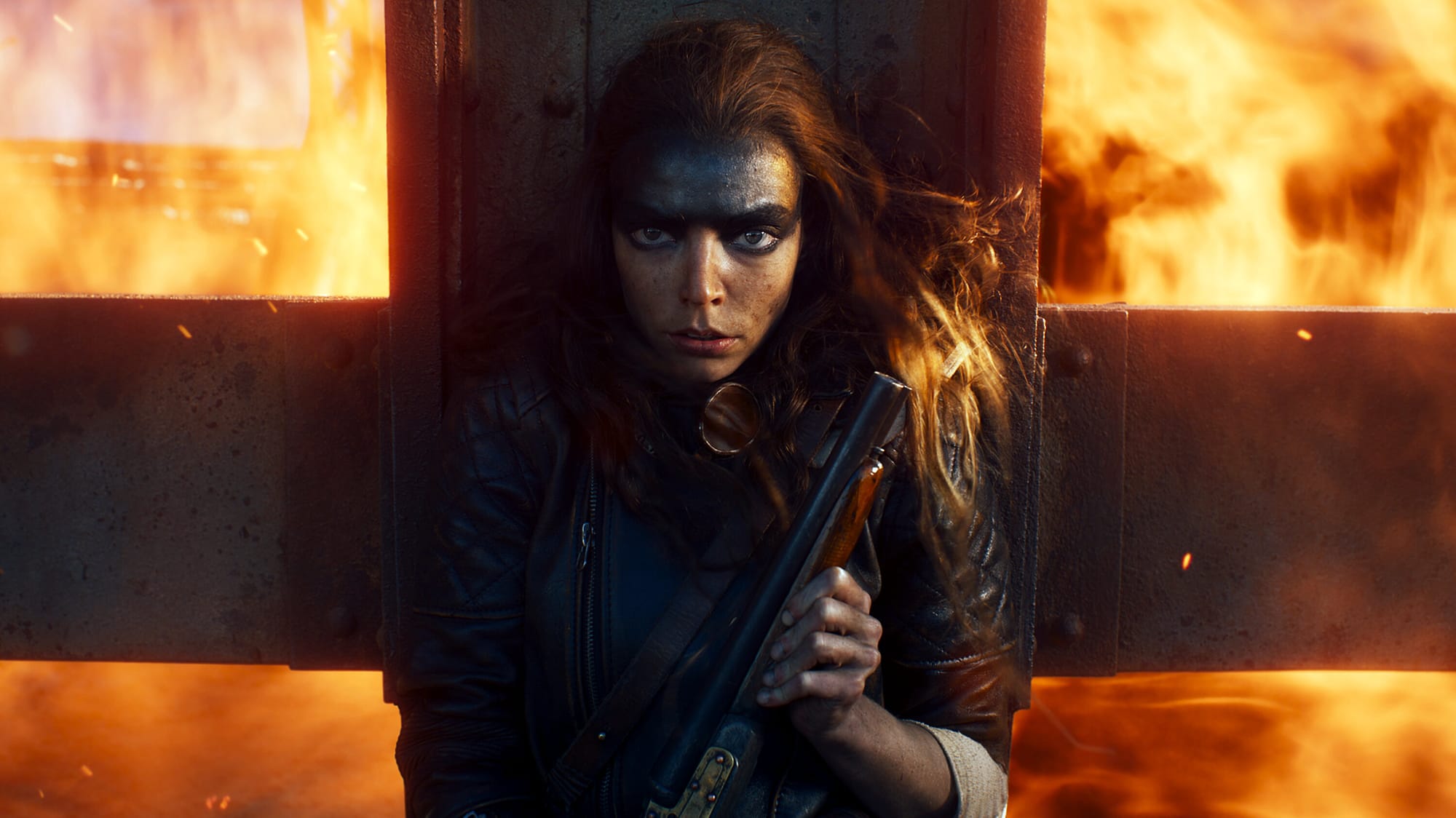 A woman with wild red hair and black grease paint over the top half of her face holds a shotgun and leans against a cross with flames raging in the background.