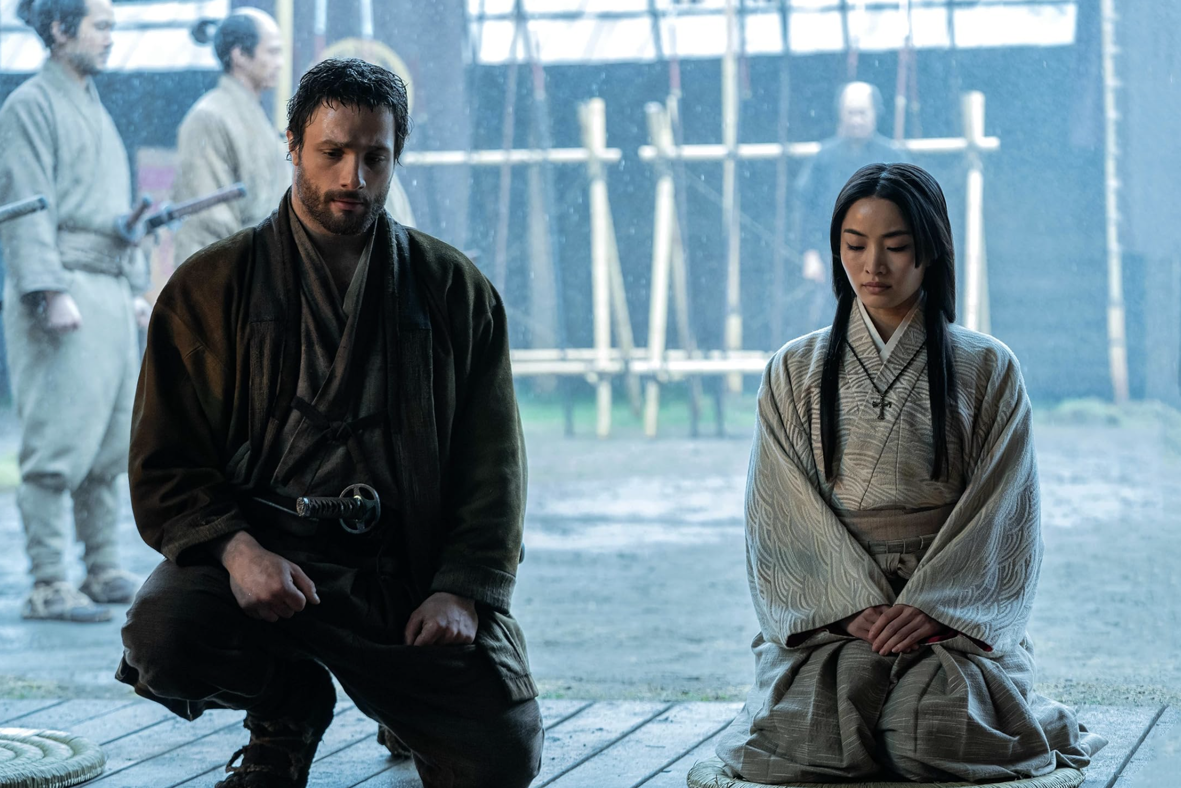 Two people kneel in a historical setting with two Japanese guards in the background. The first person is a British man wearing Japanese clothing, and the second is a Japanese woman in a kimono.