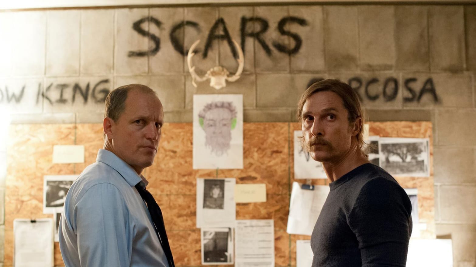 Two men stand before a crime board with the words "yellow king", "scars," and "caracosa" spray painted above the evidence.