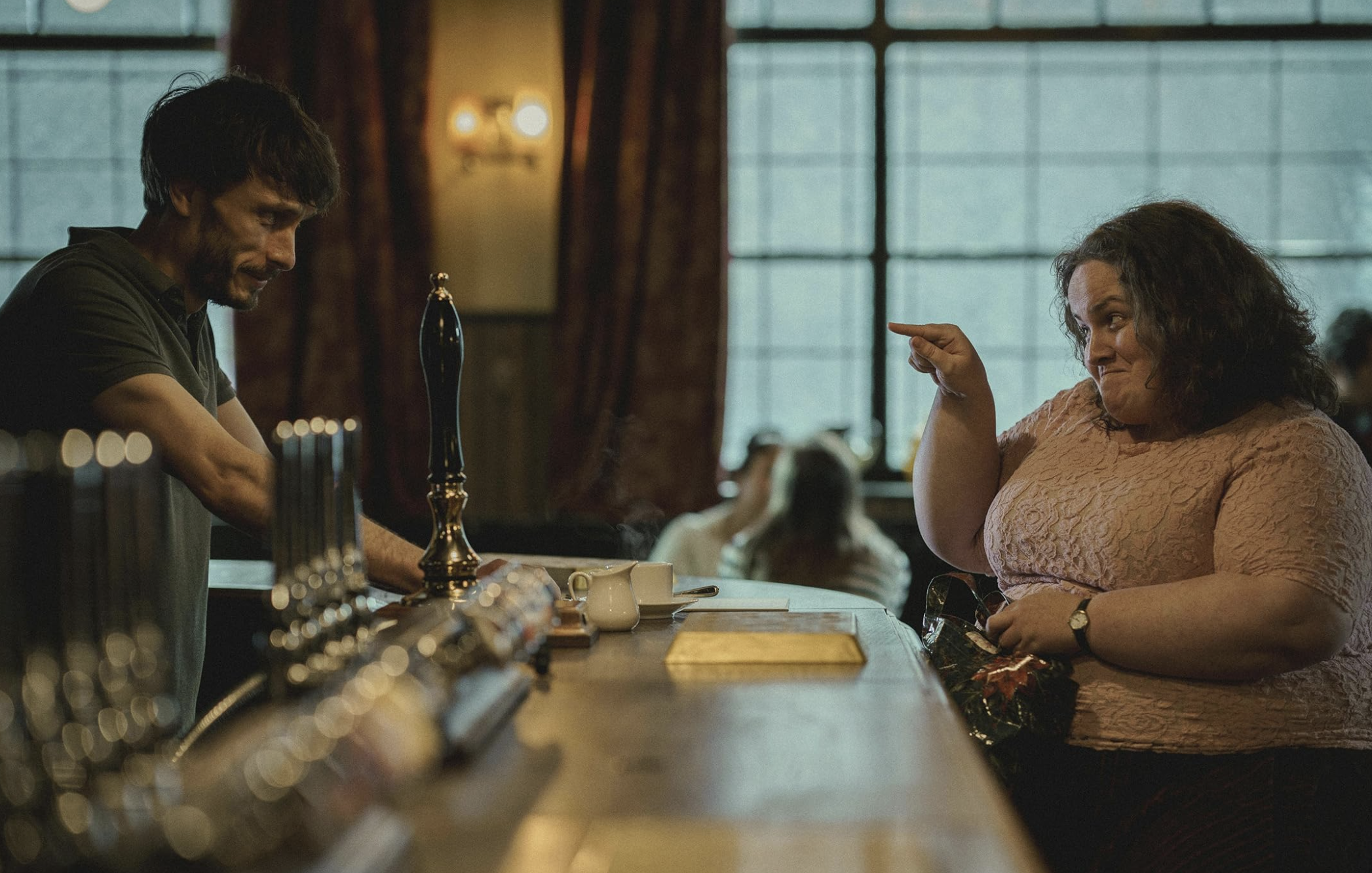 A man stands behind a bar top looking skeptically at a woman pointing back at him with cutesy expression on her face.