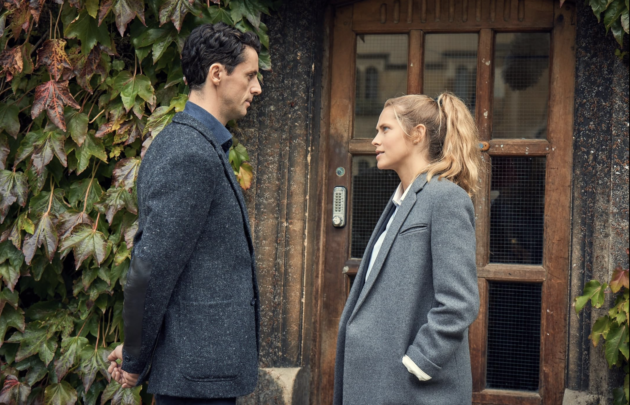 A man with dark hair in a tweed suit and a blonde woman in a blazer stare at each other with intent interest.